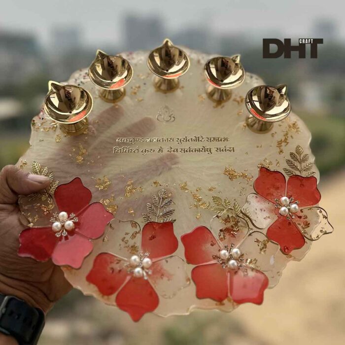 A hand holds a handcrafted Resin Aarti Thali for Pooja with, red flowers, pearls, gold leaf accents, and a Sanskrit inscription, creating a vibrant and spiritually evocative piece.