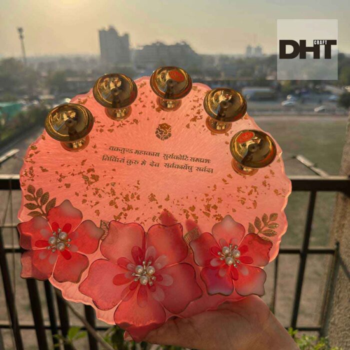 A handcrafted resin tray, adorned with five brass diyas, floral embellishments, gold leaf accents, and a Hindi inscription, is delicately held.